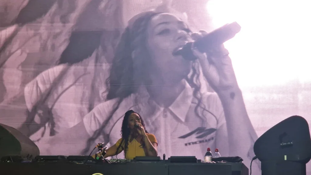 Nia Archives DJing behind a booth with a large live-feed screen of her on the microphone behind