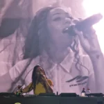 Nia Archives DJing behind a booth with a large live-feed screen of her on the microphone behind