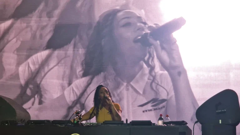 Nia Archives DJing behind a booth with a large live-feed screen of her on the microphone behind